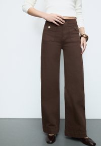 Brown wide-leg trousers made from cotton, featuring two front pockets with button accents and a clean hem. Paired with dark shoes.
