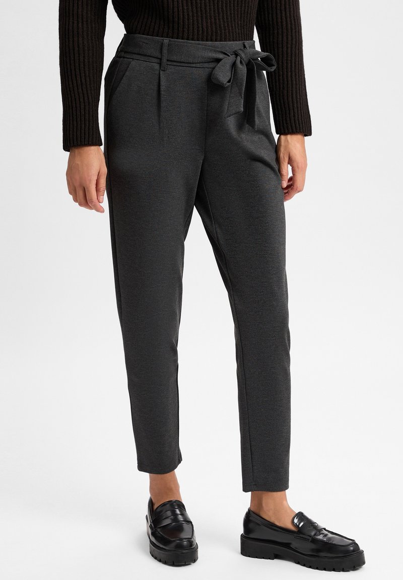 Femme portant un pantalon gris foncé fuselé avec une ceinture nouée, un pull côtelé noir, et des mocassins noirs épais, debout sur un fond blanc.