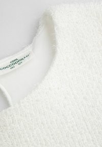 Biały top z teksturą puchatych włókien, wyposażony w gładką wewnętrzną podszewkę. Metka z napisem "Coccodrillo" wskazuje rozmiar 10Y. Okrągły dekolt.