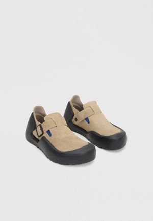 Paire de chaussures en daim beige avec semelles et brides en caoutchouc noir, avec de petits accents bleus sur un fond blanc uni.