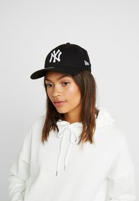 Juodas beisbolo kepurė su balta siuvinėta Niujorko "Yankees" logotipo, su išlenktu kraštu ir reguliuojamu nugaros dirželiu.