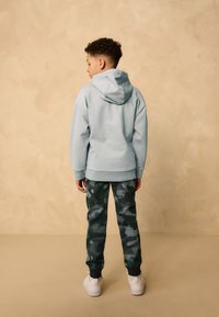 Felpa con cappuccio azzurro chiaro con tasca sul retro, abbinata a pantaloni mimetici verde scuro. Si vedono scarpe da ginnastica bianche e dettagli in tessuto strutturato.