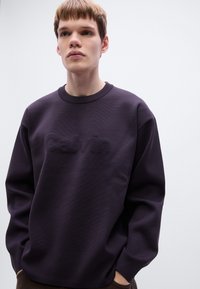 Sweatshirt violet foncé avec une finition texturée et un logo "Calvin" en relief à l'avant. Présente un col rond et des manches surdimensionnées.