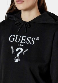 Svart hoodie med dragsko i huvan, djärv vit "GUESS®" logotyp och texten "WASHED JEANS" med grafisk detalj. Mjuk tygtextur.