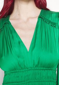 Femme aux cheveux roux portant une robe en soie vert vif avec un décolleté en V profond et des détails froncés sur les épaules et la taille.