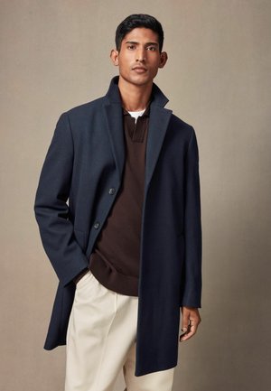 Abrigo de lana navy con cuello muesca, botones grises y bolsillos frontales, superpuesto a un suéter marrón y pantalones crema. Textura suave.