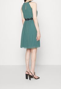 Ärmlös tealklänning med hög krage, plisserad kjol och svart blommig bältesdetalj. Ett par svarta klacksandaler synliga.