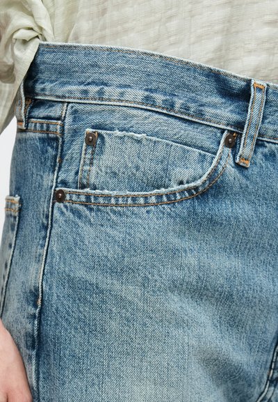 Világoskék farmernadrág fakó hatással, magas derékkal és kontrasztos narancssárga cérnavarrással. Kis elülső zsebbel és fém szegecsekkel ellátva.