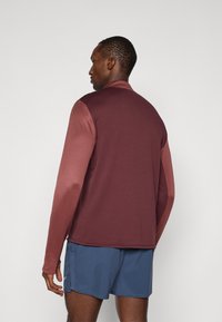 Camiseta deportiva de manga larga en burdeos oscuro con una textura suave, que presenta un diseño en bloques de color en los hombros y la parte superior de las mangas.