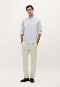 Camicia button-up a righe blu chiaro con maniche lunghe, pantaloni crema e mocassini neri. La camicia ha un piccolo logo sul petto.