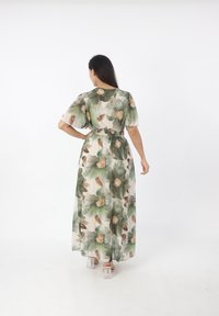 Abito maxi floreale in tessuto trasparente con foglie verdi e fiori beige, dotato di vita cinchata e maniche corte. Tacchi a blocco trasparenti.