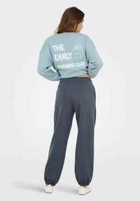 Felpa corta azzurra con testo bianco e grafica di un cartone del latte sul retro, abbinata a pantaloni da tuta larghi grigi scuri.