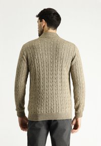 Beiger, gestrickter Pullover mit Zopfmuster, hohem Kragen, gerippten Bündchen und Saum. Verfügt über ein strukturiertes, vertikales Muster. Getragen mit grauen Hosen.