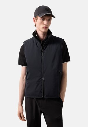 Young man wearing a black cap, short-sleeve black shirt, and a zipped black sleeveless vest with hands in pockets.