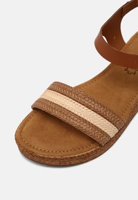 Sandales marron avec des brides tressées en tons foncés et clairs. Caractéristiques d'une semelle intérieure douce semblable au daim et d'une semelle extérieure texturée. Design plat.