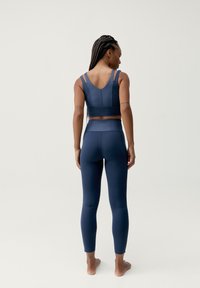 Tamsiai mėlynas sportinis komplektas, kuriame yra pritvirtinta liemenėlė su plonomis petnešomis ir derančios aukštos liemens tamprės. Lygus, elastingas audinys be raštų.