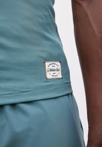 Κοντινή λήψη πετρόλ ριμπ μπλουζάκι Nike Athletic Club με ραμμένο λογότυπο και αντίστοιχα σορτς που φοράει άτομο με σκούρο χρώμα δέρματος.