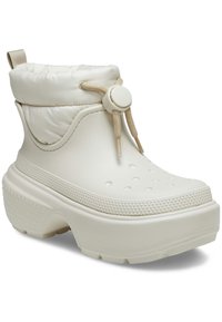 Botas tobilleras ligeras de color crema con un puño acolchado, cierre con cordón y suela texturizada que presenta pequeños agujeros y puntos elevados.