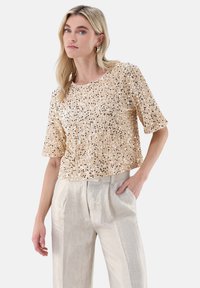 Kurz geschnittenes Oberteil mit kurzen Ärmeln in hellbeige, verziert mit verstreuten goldenen und schwarzen Pailletten. Kombiniert mit hellgrauen, hoch taillierten, plissierten Hosen.