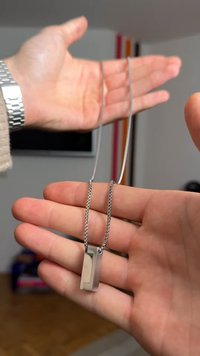 Colar com corrente prateada e pendente retangular. O pendente apresenta uma superfície polida e elegante, com um design moderno, pendurado numa corrente texturada.