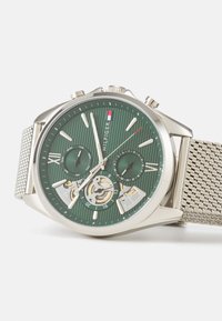 Silver mesh strap watch featuring a green striped dial, Roman and Arabic numerals, and two sub-dials with a visible movement mechanism.
