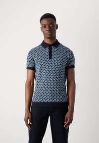 Młody mężczyzna w dopasowanej, niebieskiej koszulce polo z krótkim rękawem w wzory, z czarnym kołnierzykiem i mankietami, stojący na tle jednolitej białej ściany.