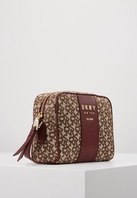 DKNY-handväska med ett mönster av bruna och krämfärgade logotyper, läderdetaljer i bourgogne, guldhardware och dragkedjestängning med en sidotassel.