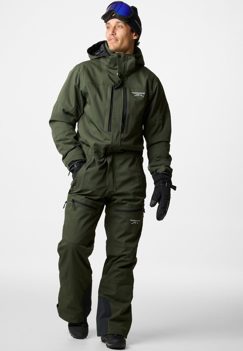 Zielony wodoodporny kombinezon narciarski z zamkiem z przodu, wieloma kieszeniami, regulowanymi mankietami i czarnym akcentem przy kostkach. Na rękach czarne rękawice i okulary ochronne.