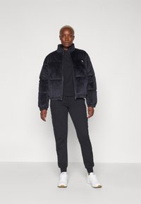 Giacca a vento imbottita in pelliccia sintetica nera con colletto alto, abbinata a pantaloni tuta neri. Sneakers bianche con suola leggera completano l'outfit.