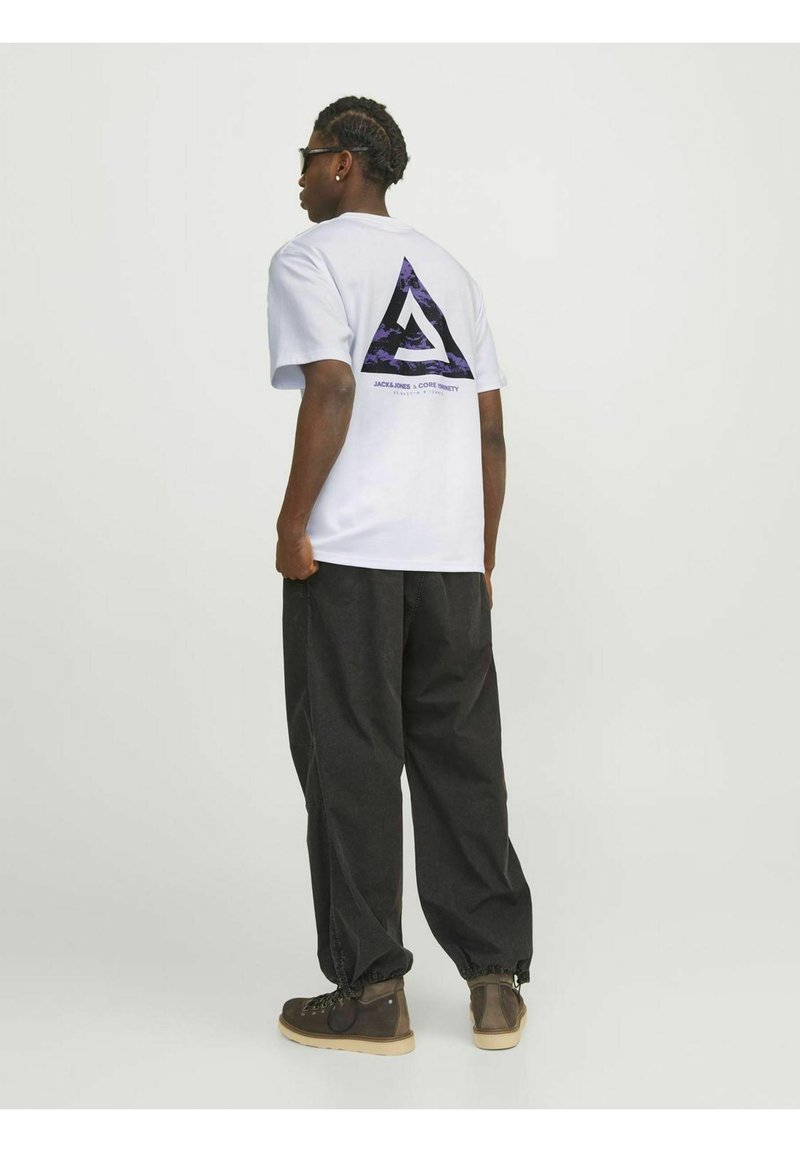 T-shirt bianco a maniche corte con un design a triangolo viola e nero sul retro, abbinato a pantaloni neri e stivali marroni.