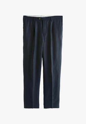 Junger Mann steht drinnen und trägt einen dunkelblauen Anzug, hellblaues Hemd, beige gemusterten Krawatte und braune Loafers vor einer beigen Wand.