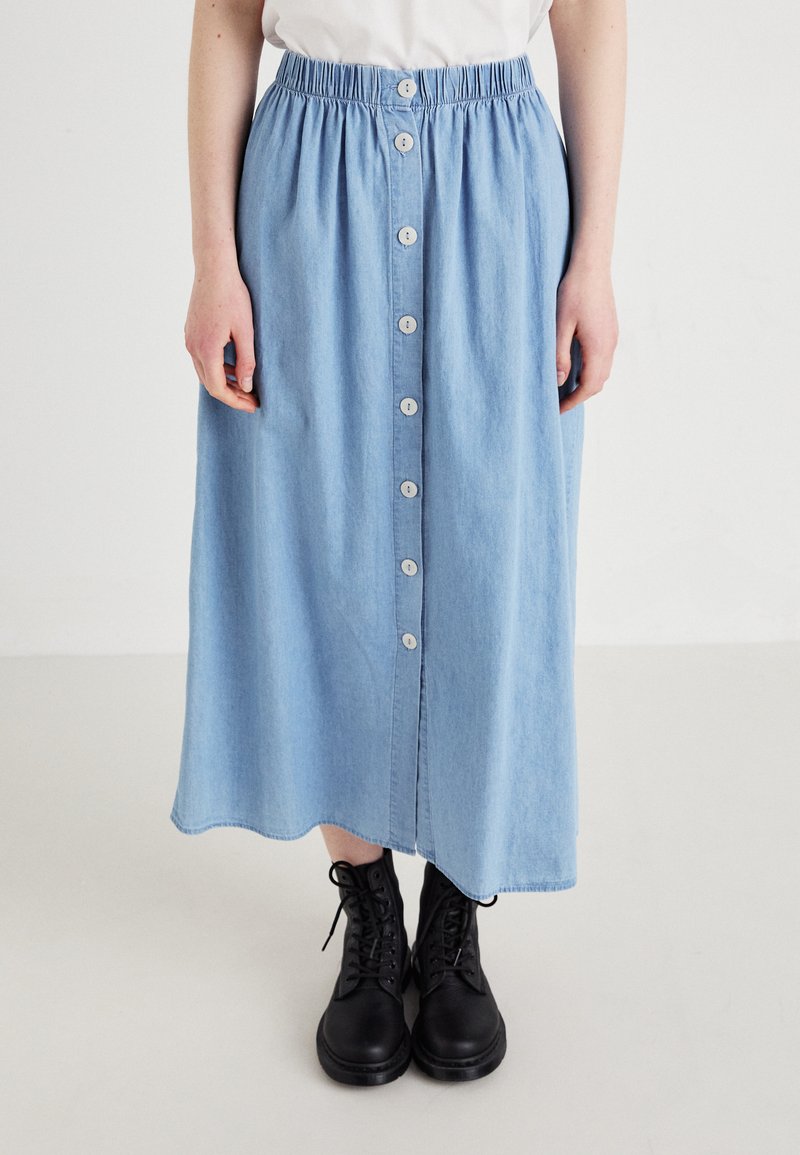 Gonna di jeans azzurri con cintura elasticizzata, caratterizzata da una chiusura frontale a bottoni e un orlo leggermente curvato, abbinata a stivali neri con lacci.