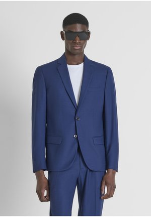 Traje azul a medida con cierre de dos botones, un bolsillo en el pecho y dos bolsillos frontales. Llevado sobre una camiseta blanca. Textura de tela suave.