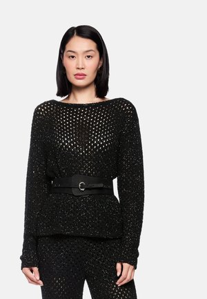 Woman with straight black hair wears a black open-knit sweater and pants set, cinched at the waist with a wide black belt against a white background.