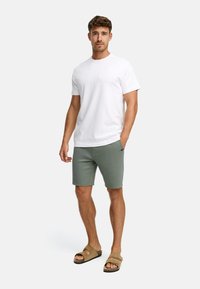 Mand iført en ensfarvet hvid t-shirt, olivengrønne shorts og brune slid-sandaler, stående foran en hvid baggrund.