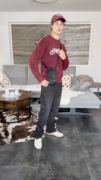 Melnas neilona crossbody soma ar regulējamu siksnu, valkāta virs bordo džempera. Modelis stāv uz tekstūras grīdas ar rakstainu paklāju zem kājām.