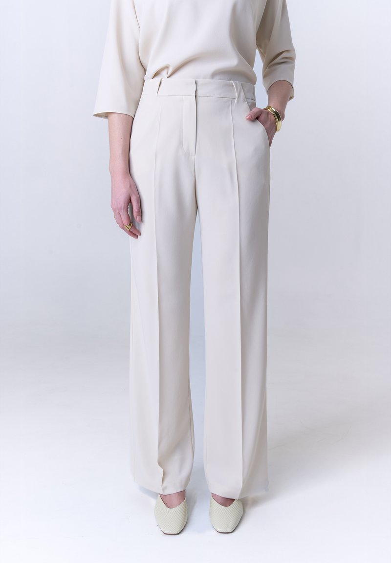 Wijde beige broeken met een soepele textuur, voorzien van plooiing aan de voorkant, zijzakken en een getailleerde fit, gecombineerd met lichte schoenen.