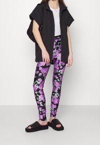 Gilet piumino nero senza maniche sopra una maglietta bianca, abbinato a leggings neri floreali con rose viola e foglie verdi, e ciabatte nere.