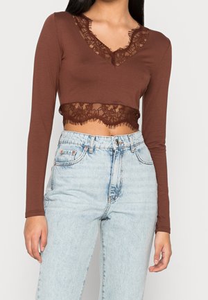 Femme portant un crop top marron à manches longues avec bordure en dentelle et un jean taille haute bleu clair sur un fond uni.