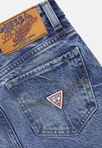 Jeans de mezclilla con un acabado azul desvaído, que presentan una etiqueta de cuero y un logo triangular rojo y blanco en el bolsillo trasero. Detalles de costura visibles.