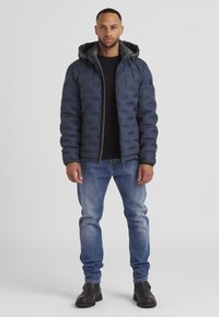 Marineblaue gep padded Jacke mit Kapuze, mit einem gesteppten Design und Reißverschluss. Getragen über einem schwarzen Shirt, kombiniert mit blauen Jeans und schwarzen Stiefeln.