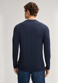 Marineblaues Langarmshirt mit rundem Halsausschnitt, glatter Textur und dezentem Branding auf der Rückseite, kombiniert mit hellblauen Jeans.