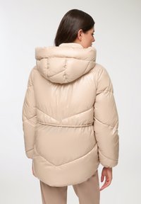 Giacca a vento beige con cappuccio, caratterizzata da sezioni trapuntate e una texture leggermente lucida. Il design include una vita cintrata e un orlo curvo.