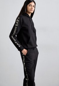 Ensemble composé d'un sweat à capuche noir et d'un pantalon en tissu doux. Le sweat à capuche possède une poche avant et un cordon de serrage ; les pantalons présentent des détails de logo sur les côtés.
