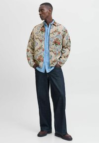 Beige shirtjack met een bloemmotief en blauwe en rode accenten, gelaagd over een lichtblauw overhemd met knopen, gecombineerd met donkere denim broek.