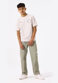 Camiseta de algodón beige claro de mangas cortas con un gráfico circular en el pecho, combinada con unos vaqueros verde claro y unas zapatillas blancas.