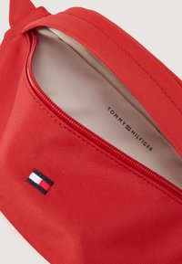 Red canvas pouch with a zipper and beige lining; features a small blue, white, and red logo patch on the front with brand name inside.