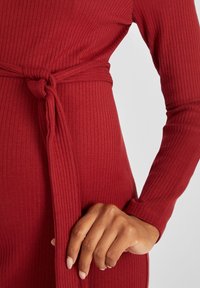 Punainen ribbikankainen mekko, jossa on pitkät hihat ja solmittava vyötäröyksityiskohta. Kangas näyttää olevan joustava ja teksturoitu, mikä täydentää istuvaa muotoilua.