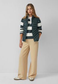 Femme portant un pantalon beige à jambres larges, une chemise rayée à manches longues, un gilet matelassé noir et des baskets, se tenant devant un fond gris clair uni.