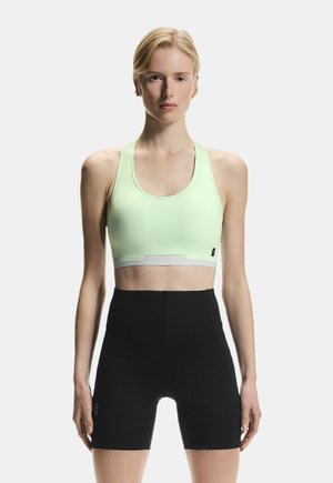 Rövid szőke hajú nő világoszöld sportmelltartóban és magasderekú fekete kerékpáros sortban áll, sima háttér előtt.